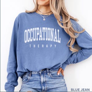 May include: A blue long-sleeve crewneck sweatshirt featuring the white text OCCUPATIONAL THERAPY printed in a collegiate-style font across the chest. The garment has a relaxed fit with ribbed cuffs and a classic round neckline design.