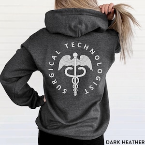 May include: Dark heather grey hooded sweatshirt with a white graphic of a medical caduceus symbol and the text "Surgical Technologist".