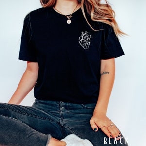 May include: Black t-shirt featuring a detailed, white anatomical heart design on the left chest. The shirt is a classic crew neck style and is paired with dark wash jeans. The word "BLACK" is printed at the bottom of the image.