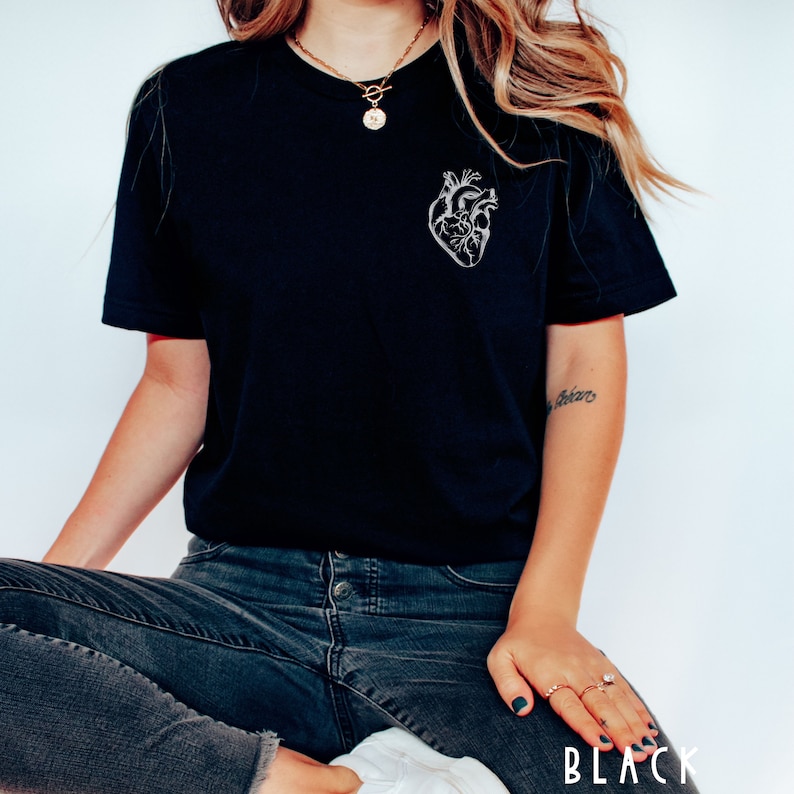 May include: Black t-shirt featuring a detailed, white anatomical heart design on the left chest. The shirt is a classic crew neck style and is paired with dark wash jeans. The word "BLACK" is printed at the bottom of the image.