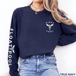 May include: A navy blue long-sleeve shirt featuring a white graphic on the left chest and down the right sleeve. The graphic depicts a caduceus symbol with the text FLOAT POOL. The shirt is paired with blue denim jeans.