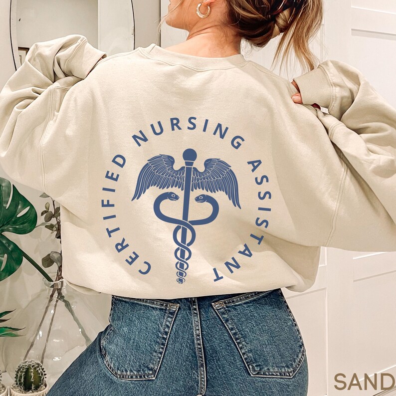 以下が含まれることがあります： 中央にカドゥケウスのシンボルが入った円の中に「CERTIFIED NURSING ASSISTANT」という文字が書かれたベージュのトレーナーです。