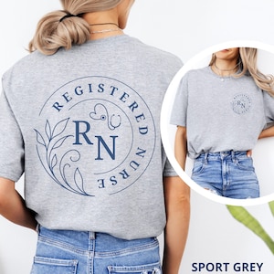 Puede incluir: Camiseta gris deportiva con un dise&ntilde;o circular azul marino en la espalda que dice "REGISTERED NURSE" alrededor de un estetoscopio, hojas y las letras "RN". Una versi&oacute;n m&aacute;s peque&ntilde;a del dise&ntilde;o est&aacute; en la parte delantera. La camiseta se combina con vaqueros azules.