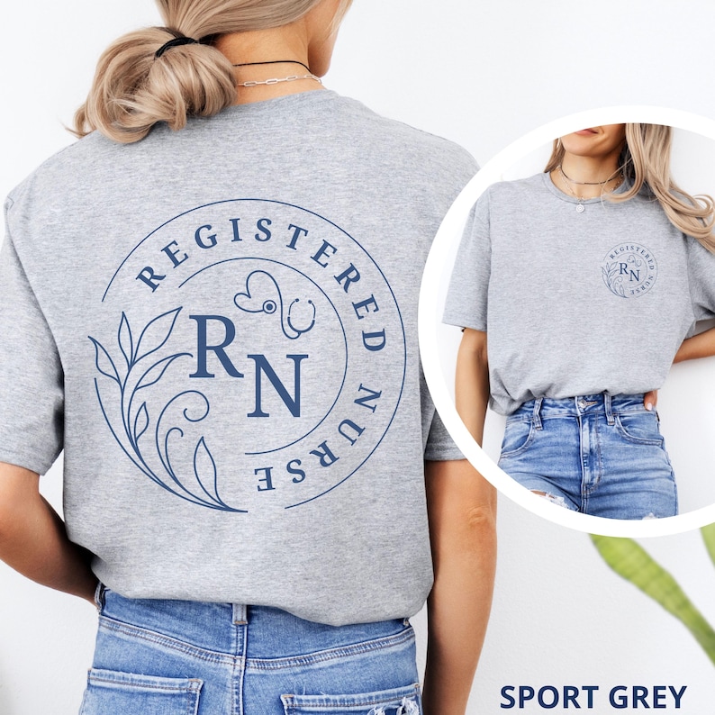 Puede incluir: Camiseta gris deportiva con un dise&ntilde;o circular azul marino en la espalda que dice "REGISTERED NURSE" alrededor de un estetoscopio, hojas y las letras "RN". Una versi&oacute;n m&aacute;s peque&ntilde;a del dise&ntilde;o est&aacute; en la parte delantera. La camiseta se combina con vaqueros azules.