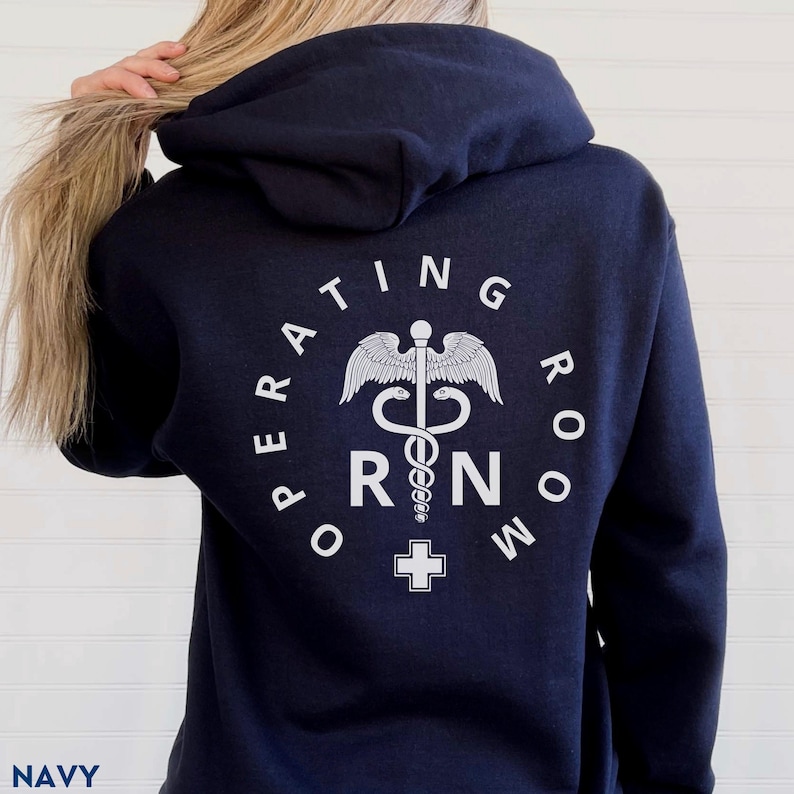 Puede incluir: Sudadera con capucha azul marino con un gr&aacute;fico blanco de un s&iacute;mbolo de caduceo m&eacute;dico dentro de un c&iacute;rculo con el texto "Operating Room RN" alrededor.
