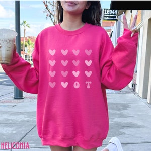 K&ouml;nnte beinhalten: Ein leuchtend pinkfarbenes Sweatshirt mit einem Muster aus rosa Herzen und den Buchstaben "VOT". Das Sweatshirt hat einen Rundhalsausschnitt und scheint aus einem weichen Material zu sein.