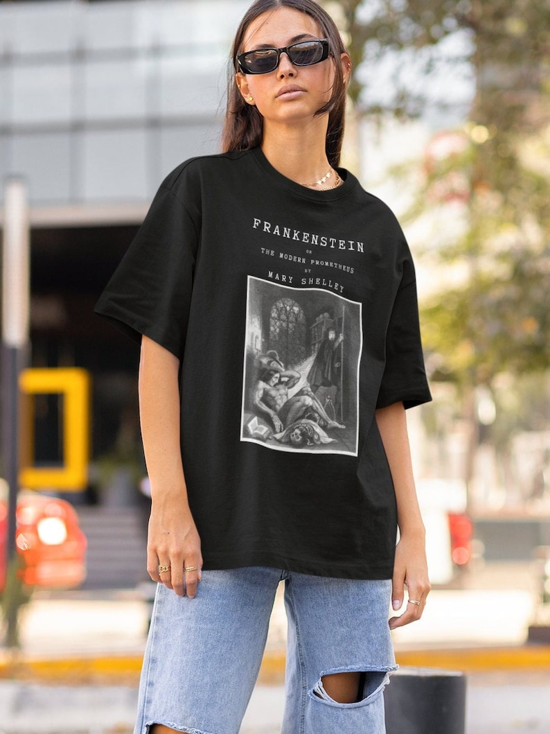 May include: Black t-shirt featuring the title "Frankenstein" and the author's name, Mary Shelley, in white text. The shirt displays a black and white illustration from the novel. The model is wearing blue jeans and sunglasses.