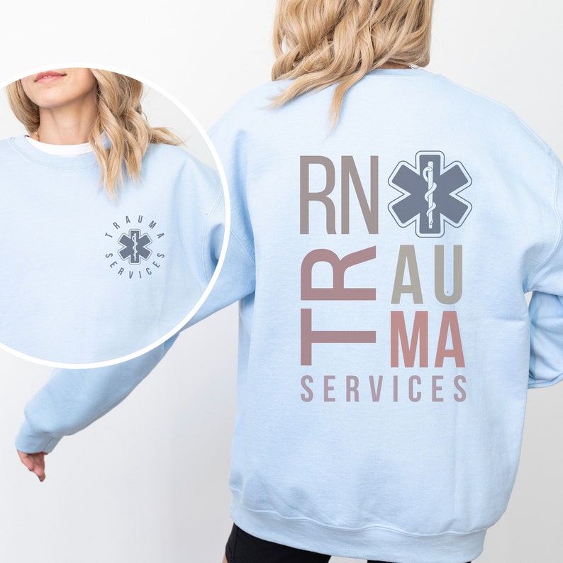 Puede incluir: Sudadera azul claro con el texto "RN" y "TRAUMA SERVICES" en rosa y gris. Una estrella m&eacute;dica gris con una serpiente est&aacute; centrada sobre el texto.