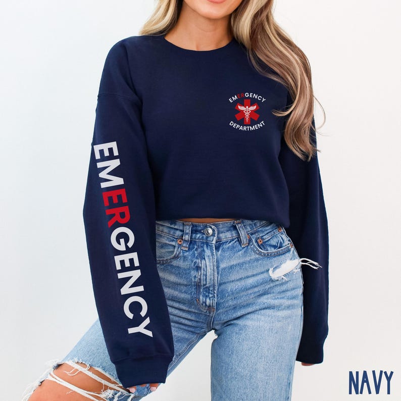 May include: A navy blue cropped sweatshirt with long sleeves. The left sleeve has the word "EMERGENCY" printed in white with a red "E". The right sleeve has a red medical symbol with a white caduceus and the text "EMERGENCY DEPARTMENT" printed in white.