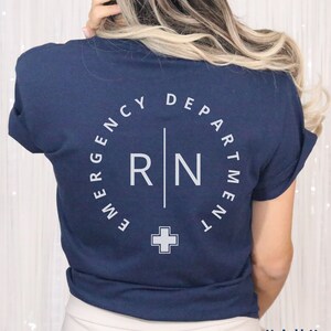 May include: Navy blue t-shirt with white text. The back of the shirt features a circular design that reads "EMERGENCY DEPARTMENT" around the top, with "RN" and a cross symbol in the center. The word "NAVY" is printed at the bottom.