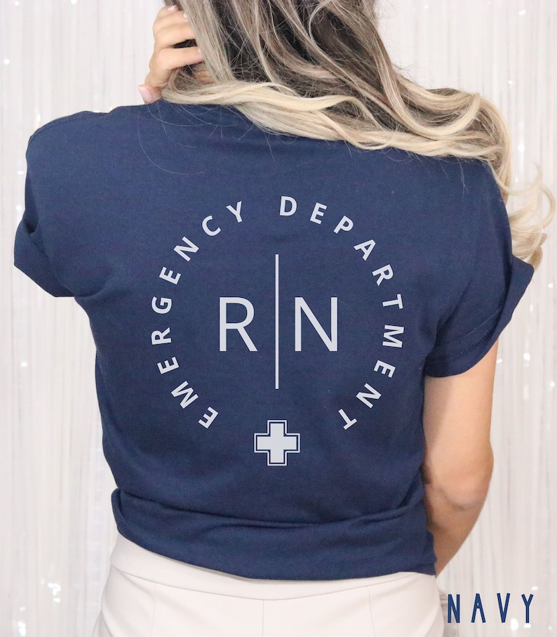 May include: Navy blue t-shirt with white text. The back of the shirt features a circular design that reads "EMERGENCY DEPARTMENT" around the top, with "RN" and a cross symbol in the center. The word "NAVY" is printed at the bottom.