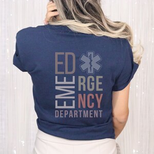 May include: A navy blue short-sleeve t-shirt featuring a graphic on the back with the text ED EMERGENCY DEPARTMENT arranged in a block layout, accompanied by a Star of Life medical symbol in a muted color palette of gray, tan, and soft pink.