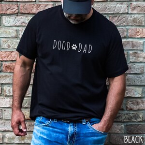 May include: Black t-shirt with the text "DOOD DAD" in a white, handwritten font, featuring a paw print in place of the "O" in "DOOD". The shirt is paired with blue jeans. The word "BLACK" is printed in white at the bottom right.