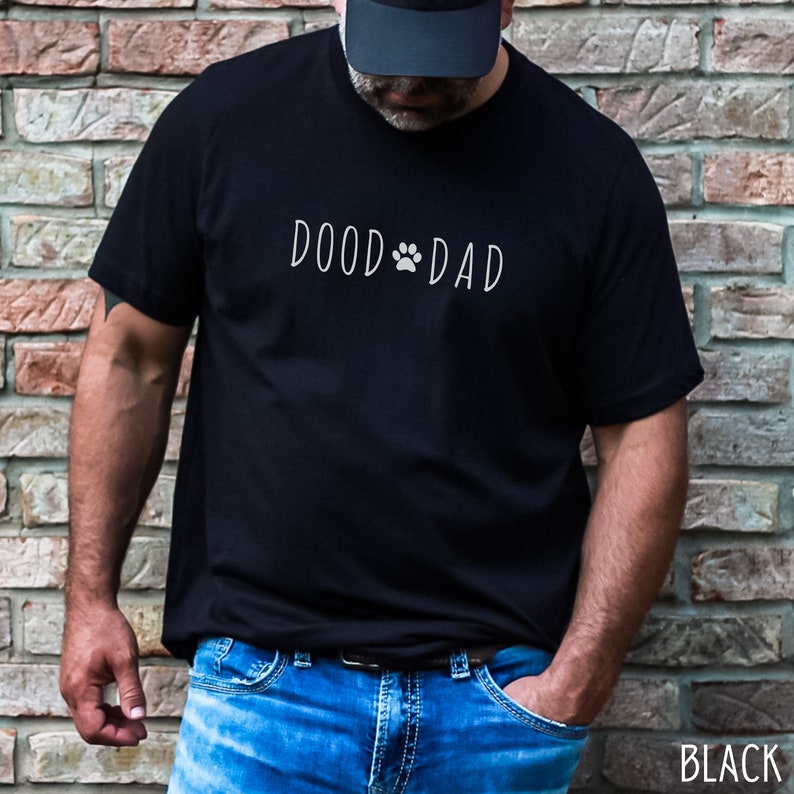 May include: Black t-shirt with the text "DOOD DAD" in a white, handwritten font, featuring a paw print in place of the "O" in "DOOD". The shirt is paired with blue jeans. The word "BLACK" is printed in white at the bottom right.