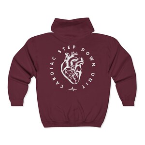 Op de afbeelding: Een bordeauxrode hoodie met ritssluiting. De achterkant heeft een witte cirkelvormige afbeelding met een anatomische hartillustratie, de woorden "CARDIAC STEP DOWN UNIT" en een hartslaglijn. De hoodie heeft een capuchon.