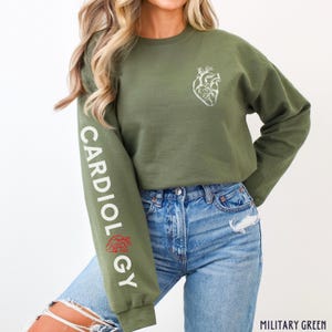 Peut inclure: Un sweat-shirt court vert &agrave; manches longues avec le mot "CARDIOLOGY" imprim&eacute; sur la manche gauche. Le sweat-shirt a un graphique de c&oelig;ur anatomique blanc sur le devant. Le mannequin porte un jean bleu.