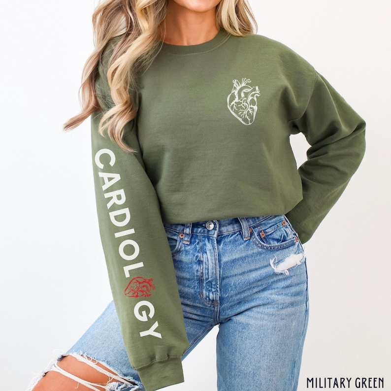 Peut inclure: Un sweat-shirt court vert &agrave; manches longues avec le mot "CARDIOLOGY" imprim&eacute; sur la manche gauche. Le sweat-shirt a un graphique de c&oelig;ur anatomique blanc sur le devant. Le mannequin porte un jean bleu.