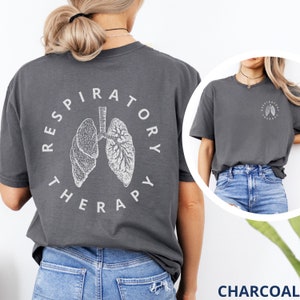 Pode incluir: T-shirt cinza carv&atilde;o com um gr&aacute;fico branco de pulm&otilde;es e as palavras "RESPIRATORY THERAPY" em um design circular nas costas. Uma vers&atilde;o menor do gr&aacute;fico est&aacute; na frente. A camisa &eacute; um estilo casual, unissex.