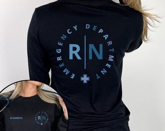 May include: A black long-sleeve shirt featuring a blue circular graphic on the back with the text EMERGENCY DEPARTMENT and RN separated by a vertical line and a small cross. The front includes the name ELIZABETH and a smaller version of the graphic.