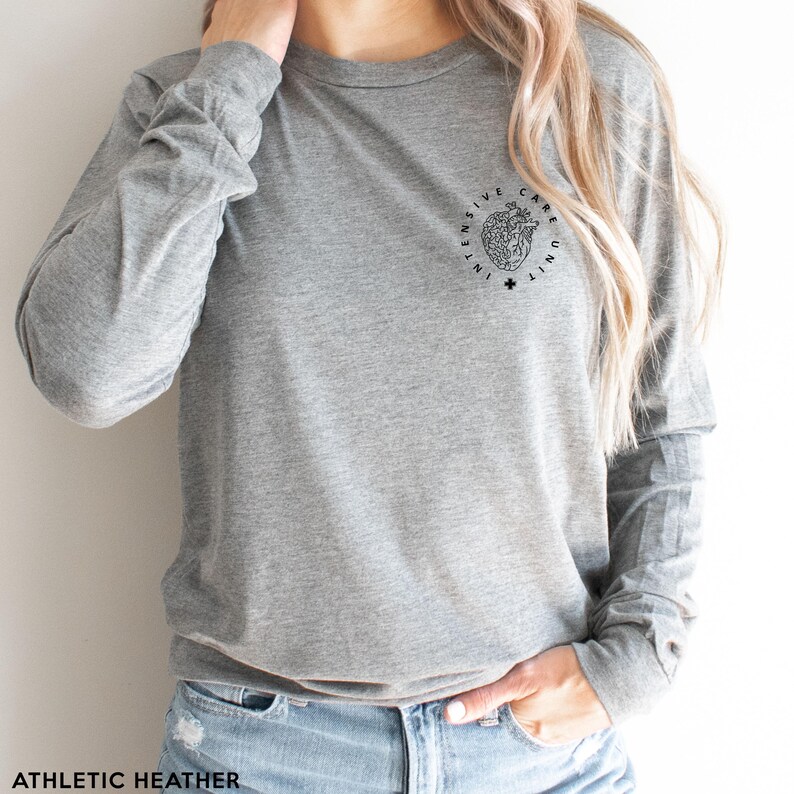 May include: Athletic heather gray long-sleeve shirt with a crew neck. The shirt features a black graphic of a heart with the words "INTENSIVE CARE UNIT" in a circle around it. A small cross is below the heart graphic. The words "ATHLETIC HEATHER" are at the bottom.