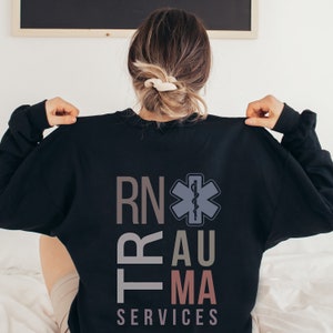 Puede incluir: Sudadera negra con el texto "RN Trauma Services" en un dise&ntilde;o vertical apilado. Un s&iacute;mbolo m&eacute;dico estilizado con una serpiente y un bast&oacute;n est&aacute; centrado sobre el texto.