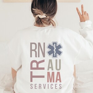 Puede incluir: Sudadera blanca con el texto "RN Trauma Services" en rosa, gris y marr&oacute;n. Un s&iacute;mbolo de estrella de la vida azul y gris est&aacute; centrado sobre el texto.
