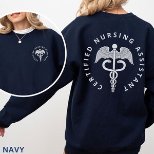 以下が含まれることがあります： ネイビーブルーのトレーナーに、白いカドゥケウスのグラフィックと「CERTIFIED NURSING ASSISTANT」の文字がプリントされています。