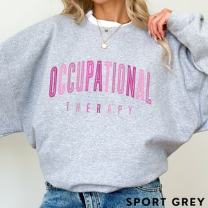 Op de afbeelding: Een grijs gem&ecirc;leerd sweatshirt met ronde hals en de tekst OCCUPATIONAL THERAPY in een gebogen, gestapeld ontwerp. De letters zijn uitgevoerd in verschillende tinten roze, van licht tot donker, met een gelaagde, omlijnde letterstijl.