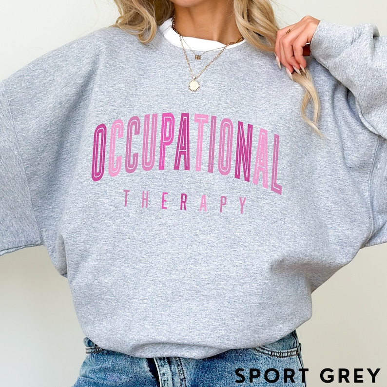 Op de afbeelding: Een grijs gem&ecirc;leerd sweatshirt met ronde hals en de tekst OCCUPATIONAL THERAPY in een gebogen, gestapeld ontwerp. De letters zijn uitgevoerd in verschillende tinten roze, van licht tot donker, met een gelaagde, omlijnde letterstijl.