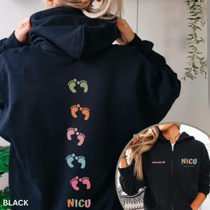 Op de afbeelding: Een zwarte hoodie met rits en een verticale rij van vijf paar babyvoetafdrukken in gedempte kleuren op de achterkant. De voorkant toont de tekst "Amanda RN" en "NICU nurse" in veelkleurige letters op de borst.
