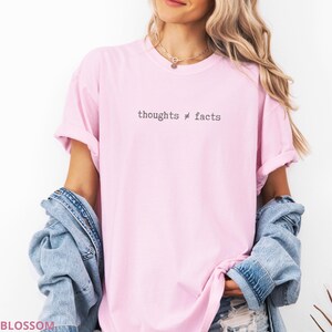 Puede incluir: Camiseta rosa claro con el texto negro "thoughts &ne; facts". La camiseta se lleva con una chaqueta vaquera. La modelo lleva un collar dorado.