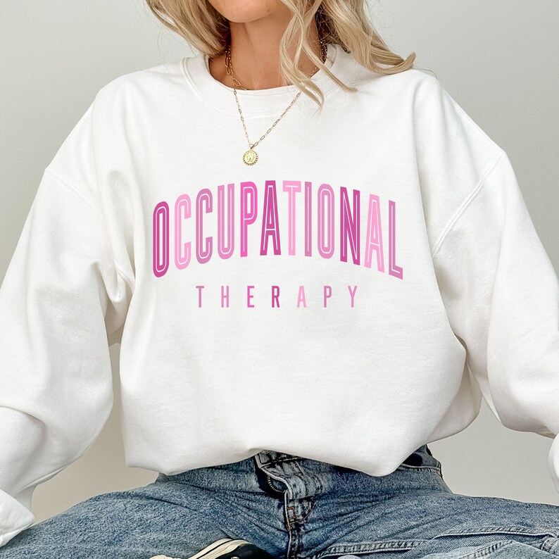 Op de afbeelding: Een wit sweatshirt met ronde hals en de tekst OCCUPATIONAL THERAPY op de borst in verschillende tinten roze. Het woord OCCUPATIONAL is gebogen in een vet, omlijnd lettertype, terwijl het woord THERAPY in een kleiner, recht lettertype staat.