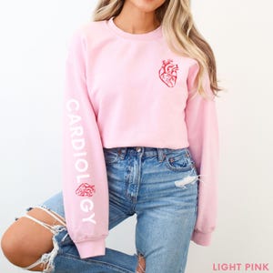 Peut inclure: Un sweat-shirt rose p&acirc;le avec le mot "CARDIOLOGY" imprim&eacute; sur la manche gauche en blanc. Un c&oelig;ur anatomique rouge est imprim&eacute; sur le devant du sweat-shirt.