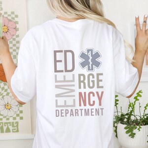May include: A white short-sleeved t-shirt featuring a graphic on the back with the text ED, RGE, NCY, and DEPARTMENT in muted tones. A Star of Life symbol is positioned next to the letters ED. The design uses soft brown, grey, and pink colors.
