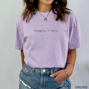 Puede incluir: Camiseta lila con la frase "thoughts &ne; facts" en una fuente sencilla. La camiseta de manga corta es de color liso. La persona lleva vaqueros azules.