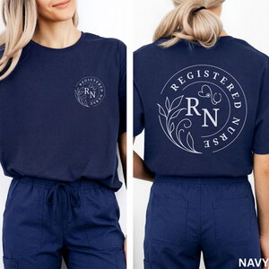 Puede incluir: Camiseta azul marino con un dise&ntilde;o blanco de enfermera titulada en la parte delantera y trasera. El dise&ntilde;o incluye las letras "RN", un estetoscopio y elementos florales. La camiseta se combina con pantalones deportivos azul marino.