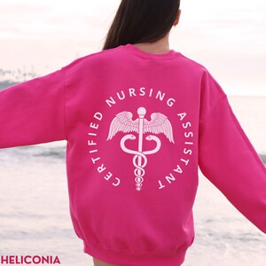 以下が含まれることがあります： 中央にカドゥケウスのシンボルが入った白い円の中に「CERTIFIED NURSING ASSISTANT」という文字が書かれたピンク色のスウェットシャツです。