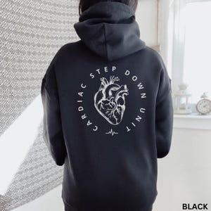 Op de afbeelding: Zwarte hoodie met een witte afbeelding op de rug. De afbeelding toont een gestileerde illustratie van een menselijk hart met de woorden "CARDIAC STEP DOWN UNIT" in een cirkelvormig ontwerp. Onder het hart bevindt zich een hartslaglijn.