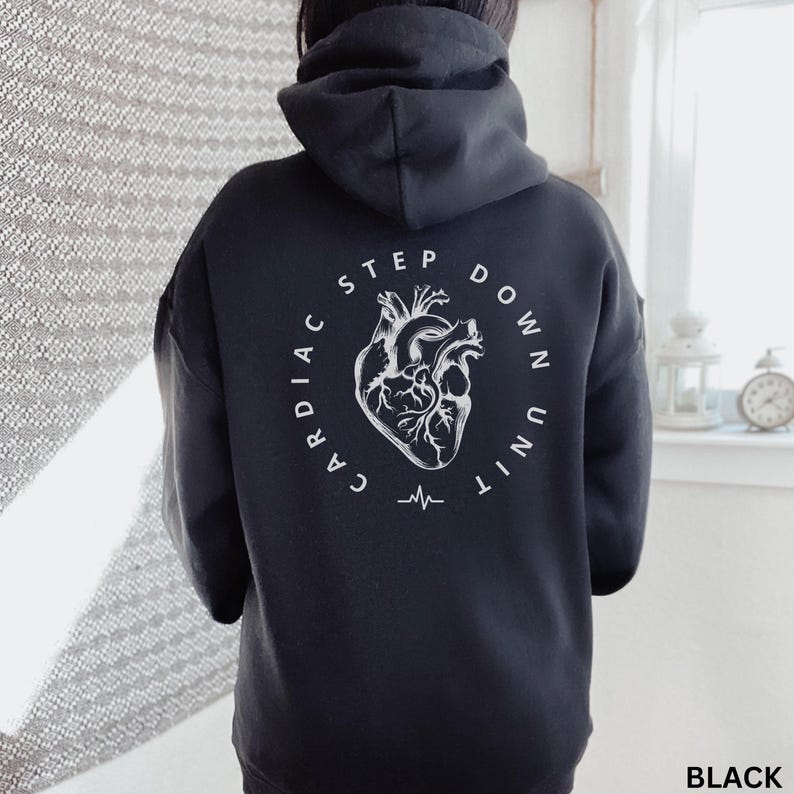 Op de afbeelding: Zwarte hoodie met een witte afbeelding op de rug. De afbeelding toont een gestileerde illustratie van een menselijk hart met de woorden "CARDIAC STEP DOWN UNIT" in een cirkelvormig ontwerp. Onder het hart bevindt zich een hartslaglijn.