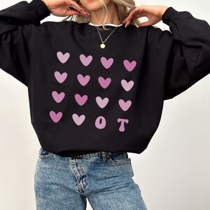 K&ouml;nnte beinhalten: Schwarzes Sweatshirt mit einem Muster aus rosa Herzen und den Buchstaben "OT". Das Oberteil hat einen Rundhalsausschnitt und lange &Auml;rmel. Das Model tr&auml;gt blaue Jeans.