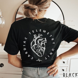 May include: A black t-shirt featuring a white graphic on the back. The design includes an anatomical heart illustration centered within a circular text arrangement that reads CARDIAC TELEMETRY UNIT, with a small heartbeat line symbol at the bottom.