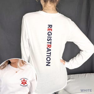 Puede incluir: Una camiseta blanca de manga larga con la palabra REGISTRATION impresa verticalmente en azul oscuro y rojo en la espalda. El pecho muestra un peque&ntilde;o s&iacute;mbolo m&eacute;dico de caduceo rojo rodeado por el texto EMERGENCY DEPARTMENT.
