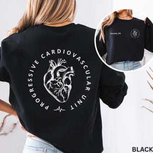 May include: A black sweatshirt featuring a white anatomical heart graphic surrounded by the text "PROGRESSIVE CARDIOVASCULAR UNIT" on the back. The front chest area displays the text "Amanda, RN" and a smaller version of the heart graphic.