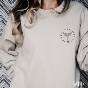 May include: A light beige sweatshirt with a black and white graphic of a medical symbol with the text "Emergency Department" around it.