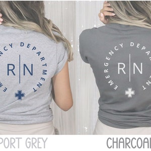 May include: Two t-shirts, one sport grey and one charcoal, featuring a circular design with the text "EMERGENCY DEPARTMENT" and "RN" in a vertical line, plus a cross symbol. The shirts are short-sleeved.