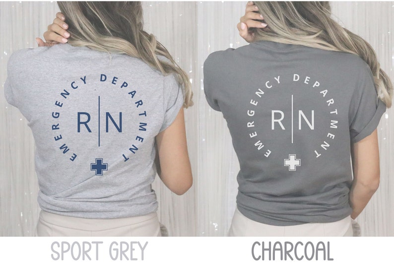 May include: Two t-shirts, one sport grey and one charcoal, featuring a circular design with the text "EMERGENCY DEPARTMENT" and "RN" in a vertical line, plus a cross symbol. The shirts are short-sleeved.