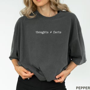 Puede incluir: Camiseta gris oscuro de gran tama&ntilde;o con el texto blanco "thoughts &ne; facts". La camiseta tiene un corte holgado y mangas cortas. La parte inferior de la camiseta es visible, junto con las manos del usuario y unos pantalones negros.