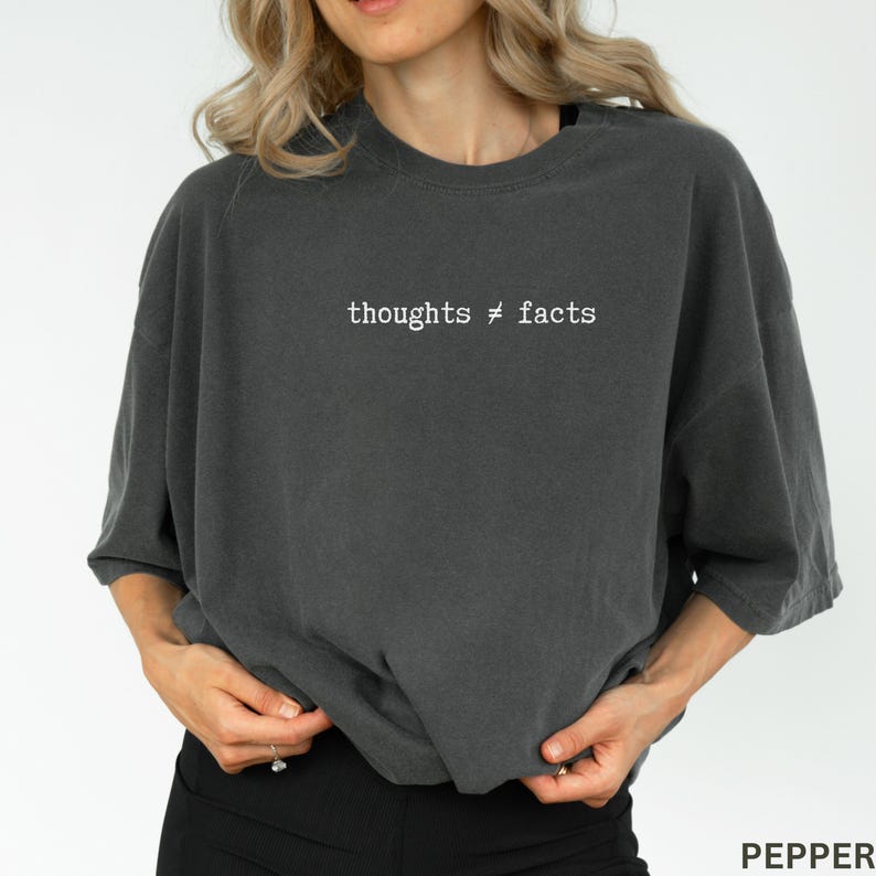 Puede incluir: Camiseta gris oscuro de gran tama&ntilde;o con el texto blanco "thoughts &ne; facts". La camiseta tiene un corte holgado y mangas cortas. La parte inferior de la camiseta es visible, junto con las manos del usuario y unos pantalones negros.