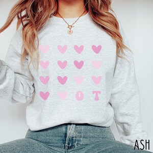 K&ouml;nnte beinhalten: Hellgraues Sweatshirt mit einem Muster aus rosa Herzen und den Buchstaben "OT" in Rosa. Das Sweatshirt besteht aus einem weichen Material und eignet sich f&uuml;r den l&auml;ssigen Gebrauch. Das Wort "ASH" ist unten rechts aufgedruckt.