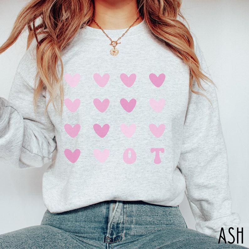 K&ouml;nnte beinhalten: Hellgraues Sweatshirt mit einem Muster aus rosa Herzen und den Buchstaben "OT" in Rosa. Das Sweatshirt besteht aus einem weichen Material und eignet sich f&uuml;r den l&auml;ssigen Gebrauch. Das Wort "ASH" ist unten rechts aufgedruckt.